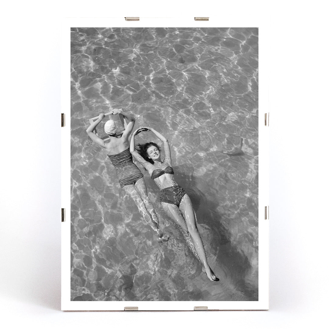 Fashion models in vintage bathing suits floating in a swimming pool artwork by Toni Frissell, framed and displayed in modern living room decor